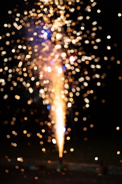 Brightly colored lights and sparks burst in a dark setting, with golden and blue bokeh effects scattered throughout the frame. The central light source appears to be a firework on the ground, creating a cascade of sparkling lights.