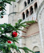 A Christmas tree branch adorned with red ornaments is in the foreground, with a historic stone building featuring arched windows in the background.