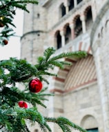 A Christmas tree branch adorned with red ornaments is in the foreground, with a historic stone building featuring arched windows in the background.