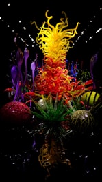 An intricate and vibrant glass sculpture featuring swirling yellow, orange, and red elements extending upwards, evoking flames. Surrounding these are ornamental green and purple glass forms resembling aquatic plants or alien flora, set against a dark background that enhances the vividness of the colors.