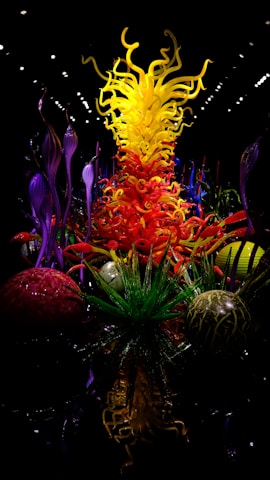 An intricate and vibrant glass sculpture featuring swirling yellow, orange, and red elements extending upwards, evoking flames. Surrounding these are ornamental green and purple glass forms resembling aquatic plants or alien flora, set against a dark background that enhances the vividness of the colors.
