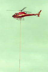 A red helicopter is flying in the sky with a long cable hanging from its underside. The helicopter has the number 286 on its side, and its rotors are in motion. The sky is a pale green color, providing a contrasting backdrop.