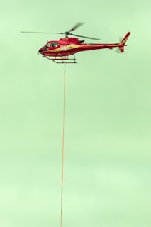 A red helicopter is flying in the sky with a long cable hanging from its underside. The helicopter has the number 286 on its side, and its rotors are in motion. The sky is a pale green color, providing a contrasting backdrop.