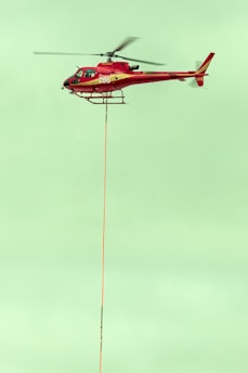 A red helicopter is flying in the sky with a long cable hanging from its underside. The helicopter has the number 286 on its side, and its rotors are in motion. The sky is a pale green color, providing a contrasting backdrop.