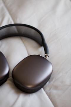 A pair of sleek, modern headphones with a metallic finish lies on a soft, off-white quilted surface. The earpads have a rounded rectangular shape, and the headband features a mesh design.