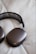 A pair of sleek, modern headphones with a metallic finish lies on a soft, off-white quilted surface. The earpads have a rounded rectangular shape, and the headband features a mesh design.