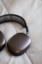 A pair of sleek, modern headphones with a metallic finish lies on a soft, off-white quilted surface. The earpads have a rounded rectangular shape, and the headband features a mesh design.