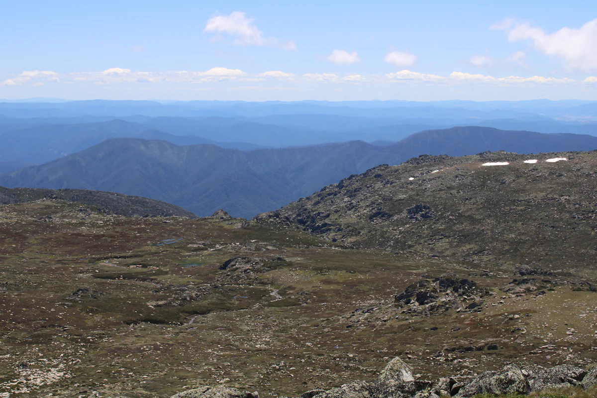 Rocky alpine grassland with a few snow patches near the top of Mt Kosciuszko, looking out over blue forested ranges