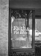 A black and white photograph of a storefront window displaying the text 'Faith & Fat Loss' in bold letters. The window reflects the silhouette of trees and a passing vehicle on the street, and blinds hang behind the glass.