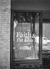 A black and white photograph of a storefront window displaying the text 'Faith & Fat Loss' in bold letters. The window reflects the silhouette of trees and a passing vehicle on the street, and blinds hang behind the glass.