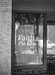A black and white photograph of a storefront window displaying the text 'Faith & Fat Loss' in bold letters. The window reflects the silhouette of trees and a passing vehicle on the street, and blinds hang behind the glass.