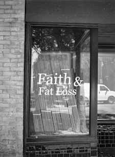 A black and white photograph of a storefront window displaying the text 'Faith & Fat Loss' in bold letters. The window reflects the silhouette of trees and a passing vehicle on the street, and blinds hang behind the glass.