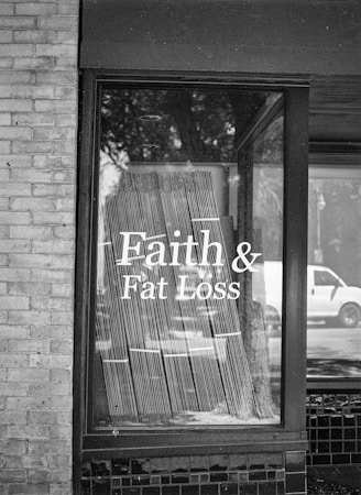 A black and white photograph of a storefront window displaying the text 'Faith & Fat Loss' in bold letters. The window reflects the silhouette of trees and a passing vehicle on the street, and blinds hang behind the glass.