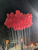 A cluster of bright red balloons is tethered together with strings, floating against a dark night sky. Warm orange lights glow from decorative street lamps below, casting shadows and adding a festive atmosphere to the scene.