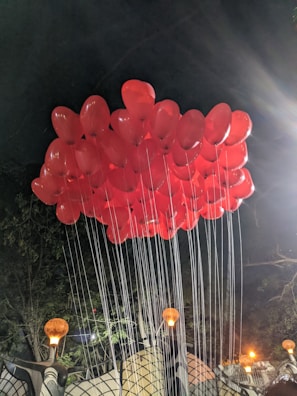 A cluster of bright red balloons is tethered together with strings, floating against a dark night sky. Warm orange lights glow from decorative street lamps below, casting shadows and adding a festive atmosphere to the scene.