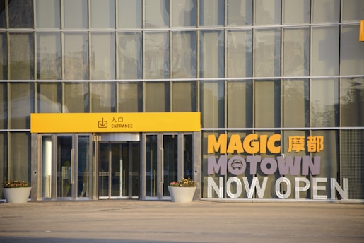 A glass facade with a prominent yellow sign marked 'Entrance' above a set of double doors. Large text on the building reads 'Magic Motown Now Open' with a mixture of bold yellow and white lettering. Two potted plants are placed symmetrically on each side of the entrance.