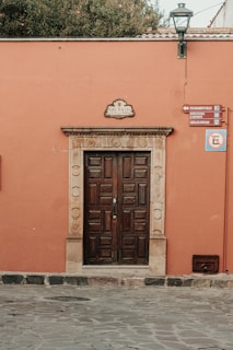 A wooden door is set within an ornate stone frame on a salmon-colored wall. A sign above the door reads 'Hdez Macias,' and several other directional signs are mounted to the right of the door. A streetlamp is attached to the wall near the top, and potted greenery is visible on top of the wall. The ground is paved with stone.