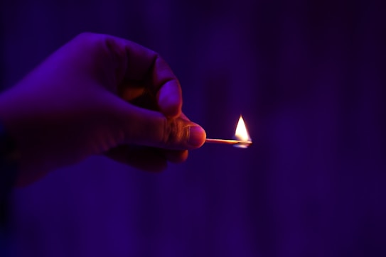 A hand holding a lit matchstick against a dark purple background. The flame on the matchstick is small and bright against the contrasting dark surroundings.