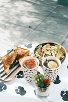 A table set for a meal with assorted food and drinks. There is a wooden tray with grilled sandwiches, a paper cup filled with iced tea accompanied by a straw, another cup with a dark-colored beverage garnished with lemon, and a glass filled with layered yogurt parfait topped with kiwi, strawberry, and blueberries. A fresh salad in a wooden bowl is next to the drinks, and the table surface has a playful black and white pattern.