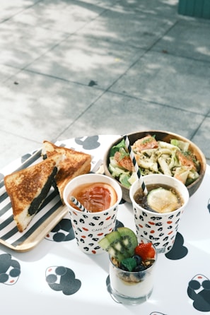A table set for a meal with assorted food and drinks. There is a wooden tray with grilled sandwiches, a paper cup filled with iced tea accompanied by a straw, another cup with a dark-colored beverage garnished with lemon, and a glass filled with layered yogurt parfait topped with kiwi, strawberry, and blueberries. A fresh salad in a wooden bowl is next to the drinks, and the table surface has a playful black and white pattern.