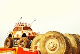A small, rusted car with large off-road tires and spikes protruding from its body, suggesting a post-apocalyptic style. The car is mounted on a platform and surrounded by several large industrial tires. The background is a pale yellow, creating a stark contrast with the rusted appearance of the car and tires.