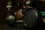 A close-up view of two high-end headphones, one with a wooden earcup and another with a matte black finish. Brand logos and model numbers are visible on each headphone, with audio equipment featuring green LED indicators in the background.