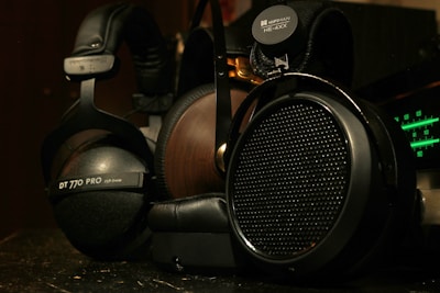 A close-up view of two high-end headphones, one with a wooden earcup and another with a matte black finish. Brand logos and model numbers are visible on each headphone, with audio equipment featuring green LED indicators in the background.