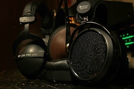 A close-up view of two high-end headphones, one with a wooden earcup and another with a matte black finish. Brand logos and model numbers are visible on each headphone, with audio equipment featuring green LED indicators in the background.