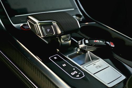 A sleek car interior featuring a gear shift with metallic and carbon fiber textures. The gear shift includes clearly labeled buttons for park, reverse, neutral, and drive. Beside it lies a car key fob with a distinctive RS logo, suggesting a high-performance model.