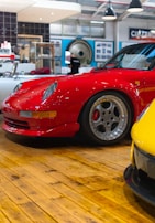 A bright red sports car is parked on a polished wooden floor within an indoor showroom. Surrounding the car, various automotive-themed decorations and memorabilia can be seen, including a black and white cinema sign and a vintage record player. Another yellow car is slightly visible in the foreground, adding to the collection.