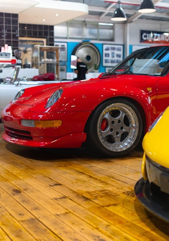 A bright red sports car is parked on a polished wooden floor within an indoor showroom. Surrounding the car, various automotive-themed decorations and memorabilia can be seen, including a black and white cinema sign and a vintage record player. Another yellow car is slightly visible in the foreground, adding to the collection.