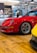 A bright red sports car is parked on a polished wooden floor within an indoor showroom. Surrounding the car, various automotive-themed decorations and memorabilia can be seen, including a black and white cinema sign and a vintage record player. Another yellow car is slightly visible in the foreground, adding to the collection.