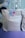A gift bag with a gray and white pattern featuring star-like shapes, topped with a shiny silver ribbon. There is a circular tag with a pink cartoon shark and the words 'Thanks Baby Shark Party'. The background includes a blurred view of balloons in shades of blue and white.