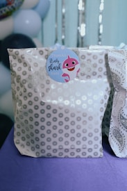 A gift bag with a gray and white pattern featuring star-like shapes, topped with a shiny silver ribbon. There is a circular tag with a pink cartoon shark and the words 'Thanks Baby Shark Party'. The background includes a blurred view of balloons in shades of blue and white.