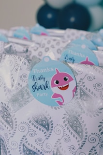 Gift bags decorated with a paisley pattern and a tag featuring a cartoon pink shark, with the words 'Thanks Baby Shark Party'. Blue and white balloons are blurred in the background.