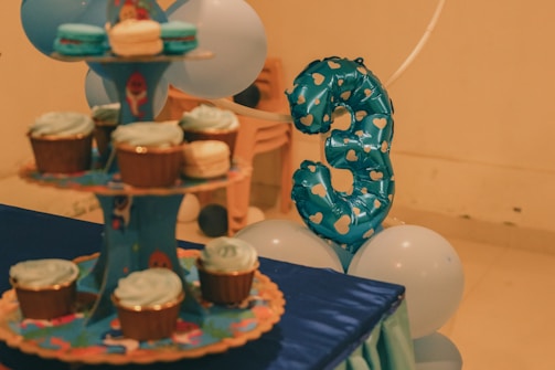 A festive setup featuring a blue balloon shaped as the number three with heart patterns. Nearby, there's a cupcake stand holding several cupcakes with light blue frosting. Blue and white balloons surround the arrangement. The setting seems to be indoors with soft lighting.