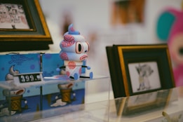 A whimsical toy figure resembling an ice cream cone with anthropomorphic features sits on a display platform. It is surrounded by brightly colored packaging and framed artwork in the background. The setting appears to be a store or gallery, evident from the price tag displayed prominently next to the toy.
