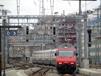 A red and gray train travels down a track surrounded by complex rail infrastructure. Overhead wires and signal lights are visible, along with a red building in the background. Several tracks and switches are present, indicating a busy rail junction or station.