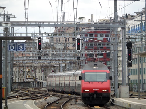 A red and gray train travels down a track surrounded by complex rail infrastructure. Overhead wires and signal lights are visible, along with a red building in the background. Several tracks and switches are present, indicating a busy rail junction or station.