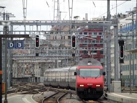A red and gray train travels down a track surrounded by complex rail infrastructure. Overhead wires and signal lights are visible, along with a red building in the background. Several tracks and switches are present, indicating a busy rail junction or station.