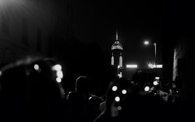 A dark urban scene at night with a tall, illuminated skyscraper in the background. The foreground features several silhouetted figures, possibly onlookers, with some bokeh light effects from street lighting or camera glare.