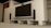 A modern home entertainment setup featuring a large flat-screen TV mounted on a light-colored wall. Two tall black speakers stand on either side of the TV. The room has a minimalist design with a clean, uncluttered look. Shelving and a wooden door are visible in the background.
