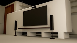 A modern home entertainment setup featuring a large flat-screen TV mounted on a light-colored wall. Two tall black speakers stand on either side of the TV. The room has a minimalist design with a clean, uncluttered look. Shelving and a wooden door are visible in the background.