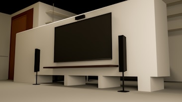 A modern home entertainment setup featuring a large flat-screen TV mounted on a light-colored wall. Two tall black speakers stand on either side of the TV. The room has a minimalist design with a clean, uncluttered look. Shelving and a wooden door are visible in the background.