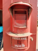 A vintage red postal mailbox with a slot for letters and a circular time plaque. The lower section features white text in a script, indicating the collection time.