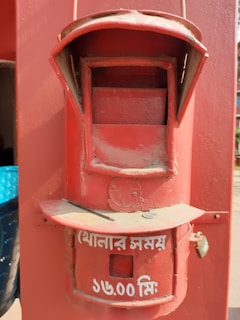 A vintage red postal mailbox with a slot for letters and a circular time plaque. The lower section features white text in a script, indicating the collection time.