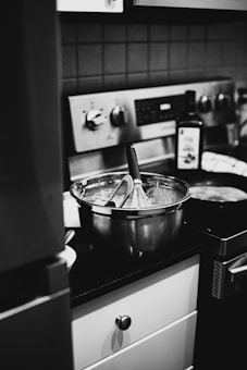 A kitchen scene featuring a metal mixing bowl on a countertop with a whisk inside, surrounded by cooking utensils and a bottle. The background shows a stove with knobs and a towel hanging nearby, all in black and white.