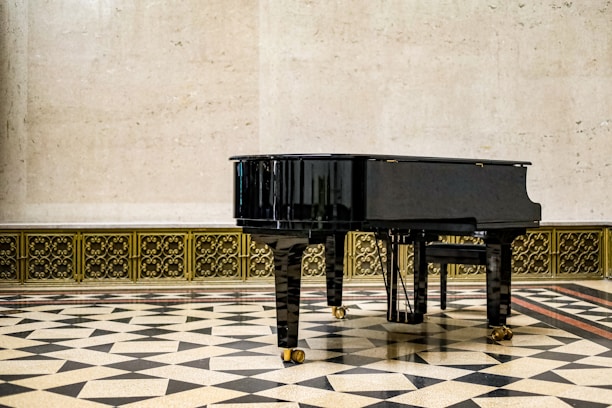 A black grand piano stands prominently on a patterned tiled floor, set against a light-colored wall with decorative metalwork along the base. The setting suggests a formal and elegant environment.