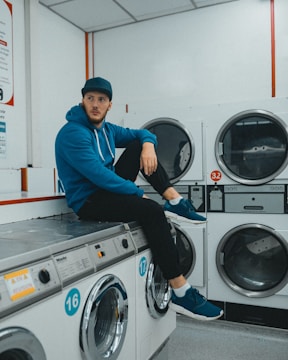 A person wearing a blue hoodie and cap sitting on a washing machine in a laundromat. The setting includes multiple washing and drying machines with numbered labels. The person appears to be in a relaxed pose.
