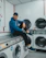 A smiling technician in a blue uniform repairing a washing machine in a bright, clean home laundry room.